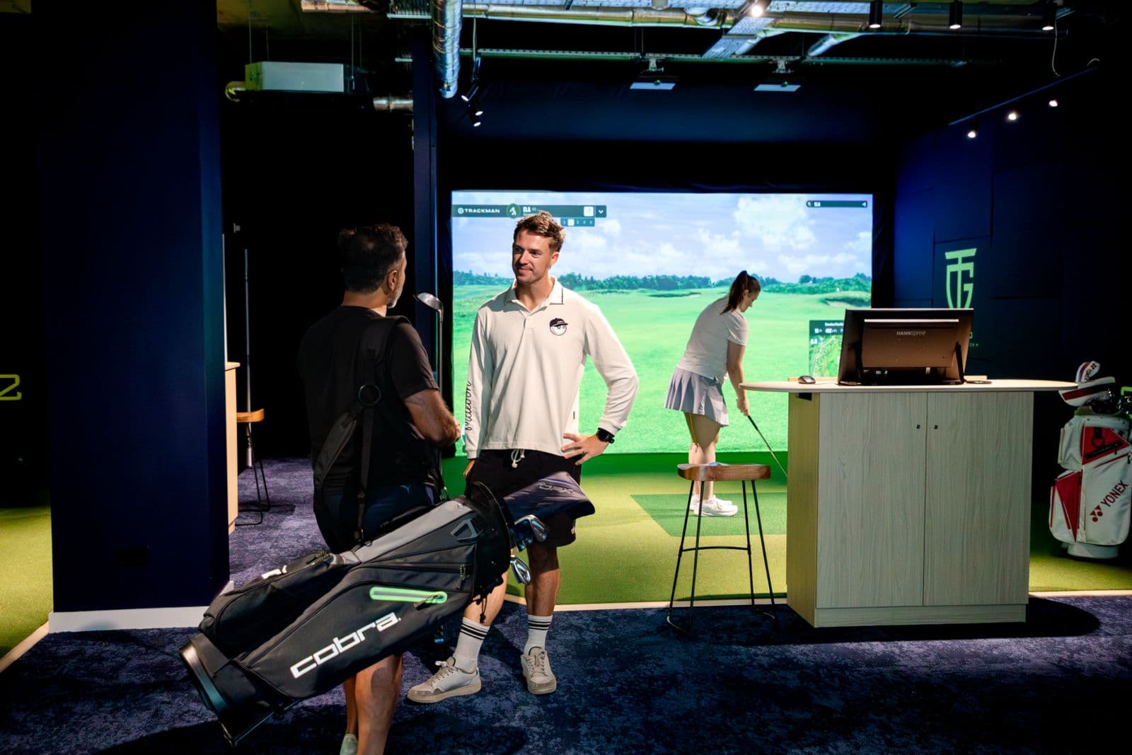 Two golfers talking in the foreground of a TeeGo simulator venue while a third plays a shot into an impact screen behind them