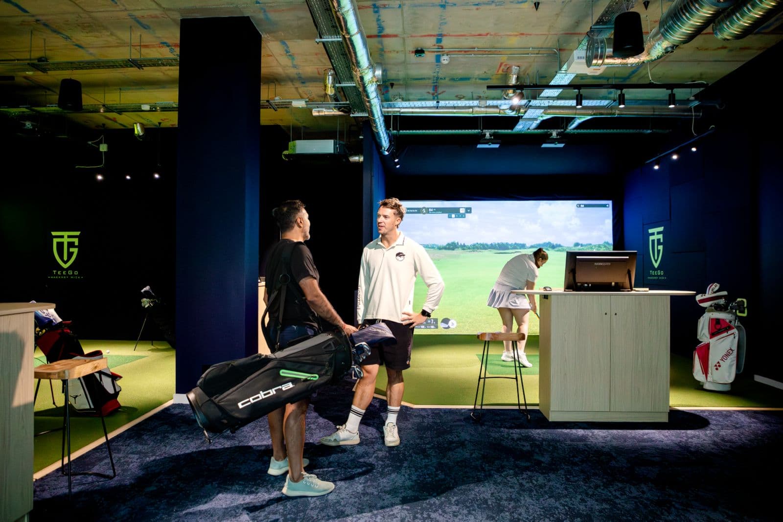 A TeeGo simulator venue: a bay with a taut impact screen showing a projected course, TeeGo branding on the walls either side, and players in the foreground